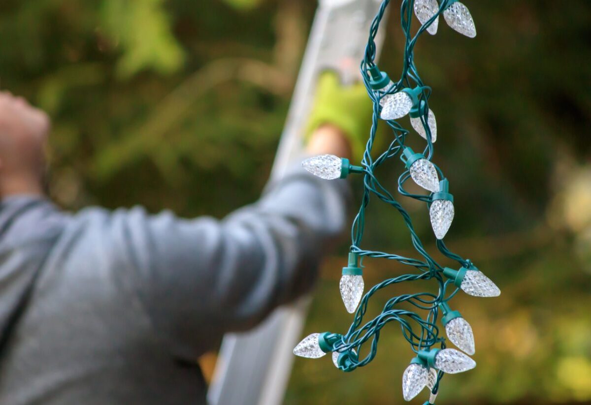 Homeowner in the process of scaling a ladder while holding a bundle of Christmas lights.