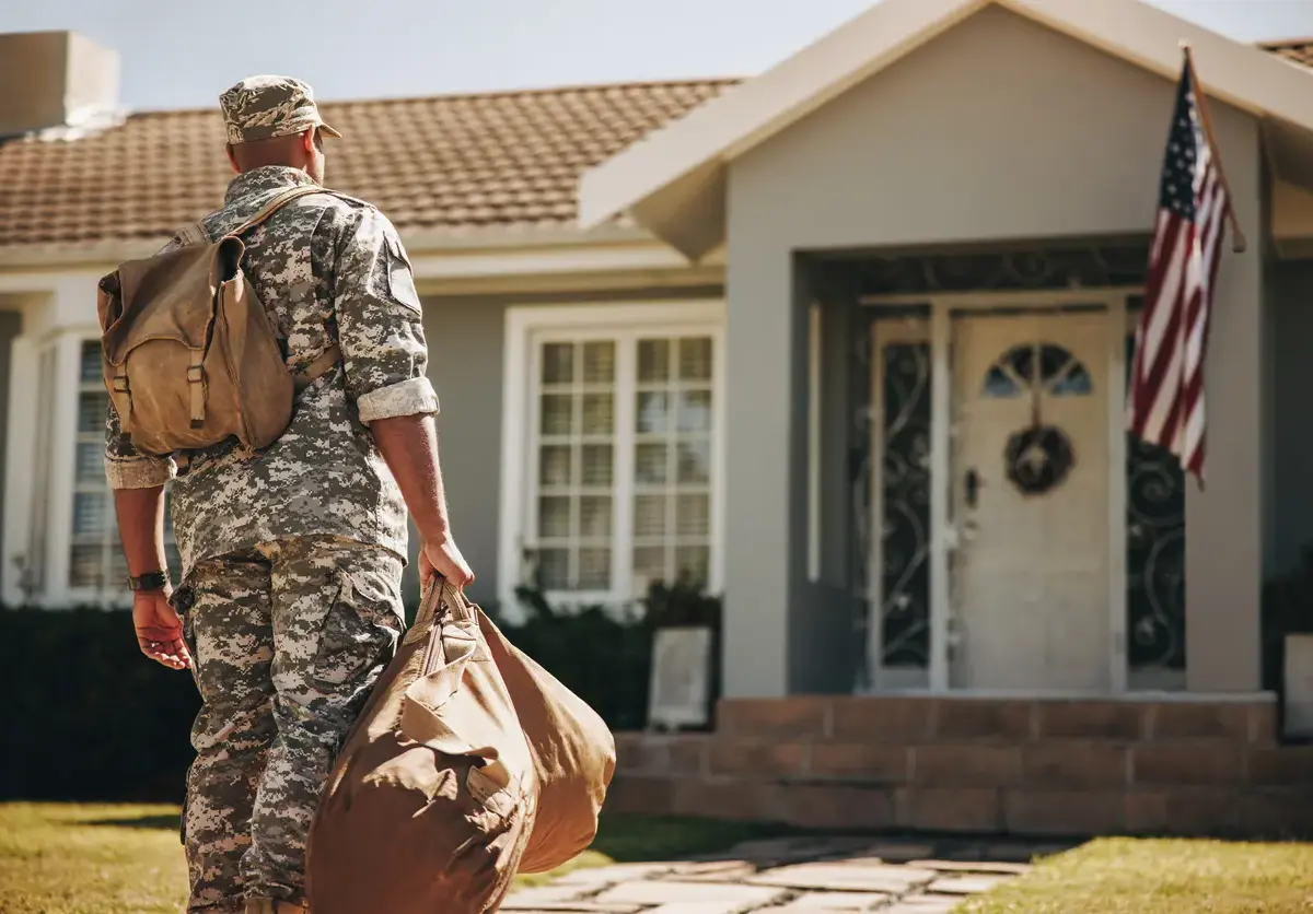 Military member looking toward the front of a home with a bag in his hand and another on his back.