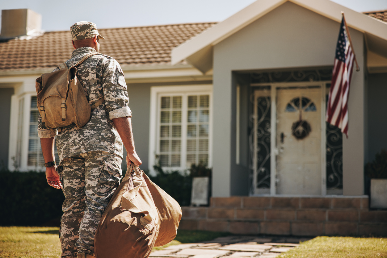 Military member looking toward the front of a home with a bag in his hand and another on his back.