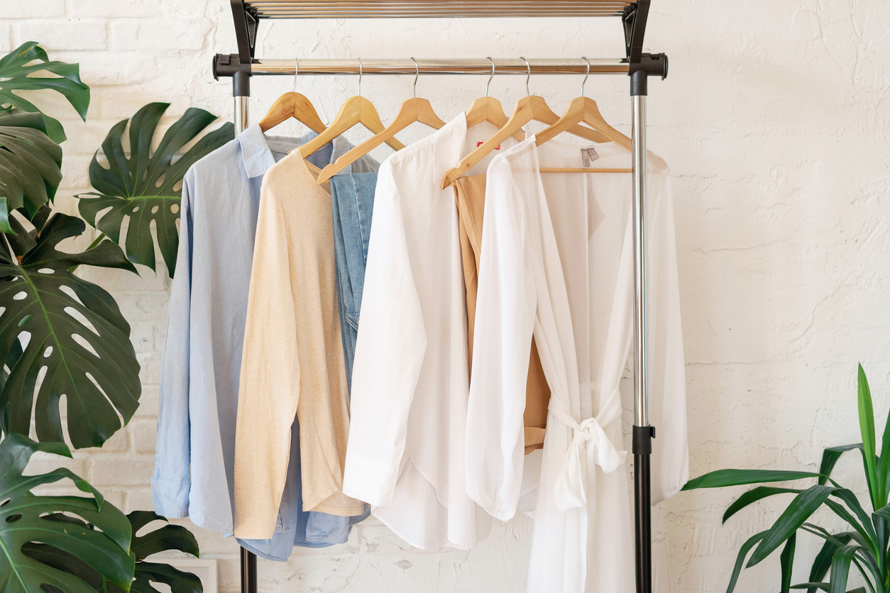 A trendy beige and light blue capsule wardrobe is displayed on a rail rack against a white-textured wall with Monstera leaves on the left.