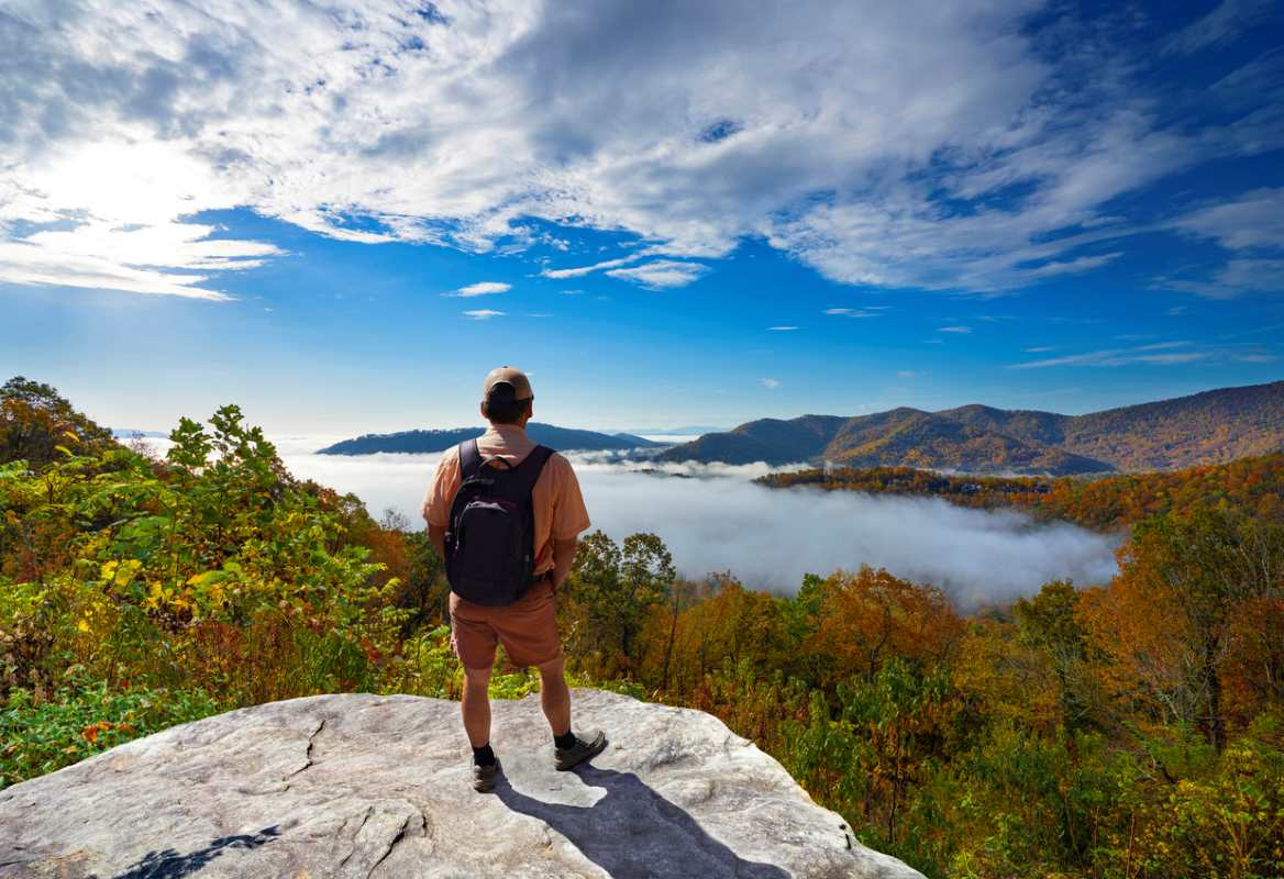 A man stands overlooking the Blue Ridge Mountains forest from a cliff on a sunny day.