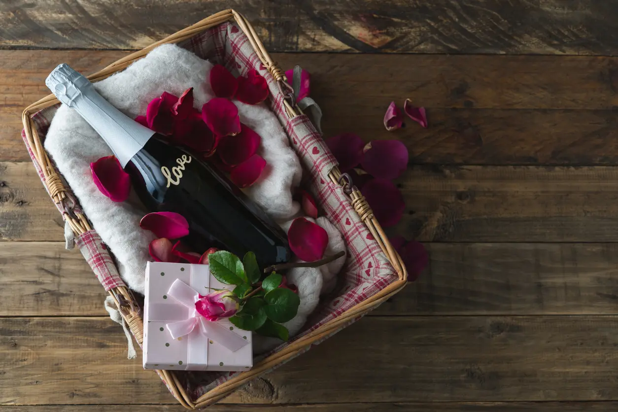 A Valentine’s Day basket with a bottle of wine, a pink present, a rose, and rose petals. 